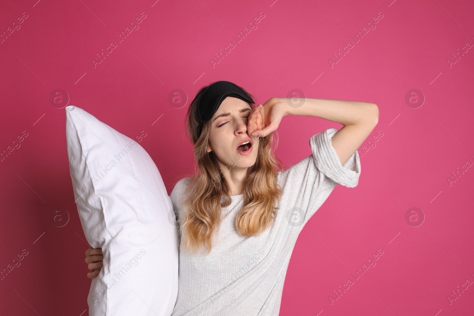 Photo of Young tired woman with sleeping mask and pillow yawning on pink background