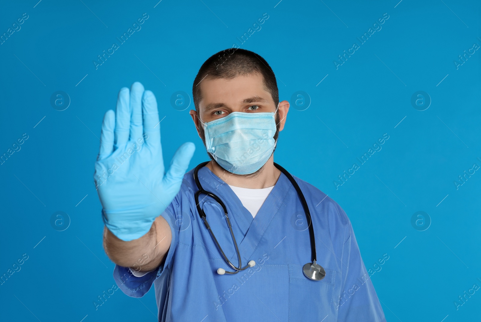 Photo of Doctor in protective mask showing stop gesture on light blue background. Prevent spreading of coronavirus