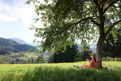 Photo of Young woman reading book under tree on meadow in mountains