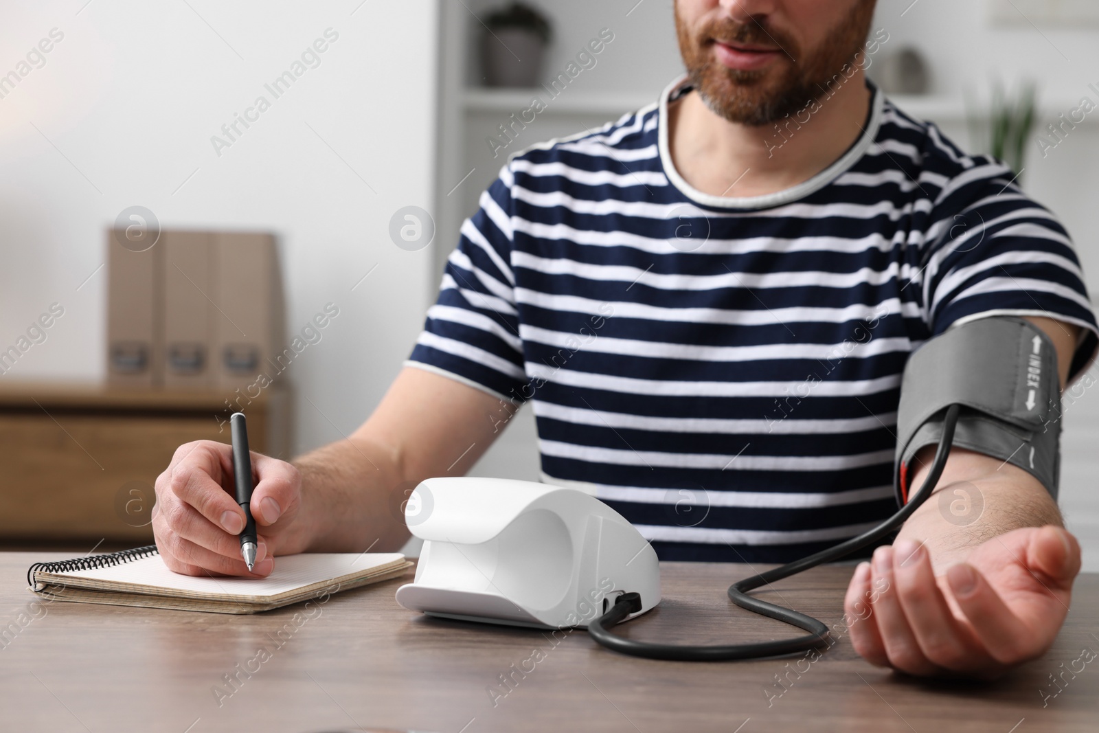 Photo of Man measuring blood pressure and writing it down into notebook in room, closeup