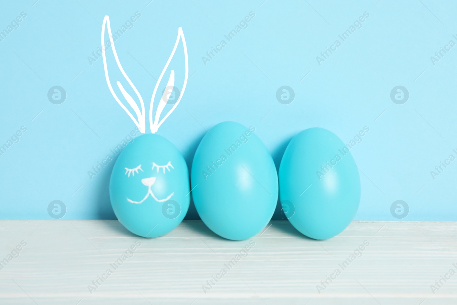 Image of One egg with drawn face and ears as Easter bunny among others on white wooden table against light blue background