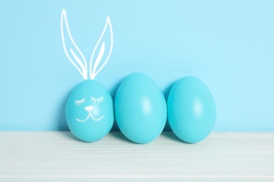 Image of One egg with drawn face and ears as Easter bunny among others on white wooden table against light blue background