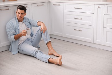 Photo of Man with cup of drink sitting on warm floor in kitchen, space for text. Heating system