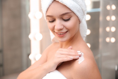 Beautiful woman with towel on head applying body cream in bathroom