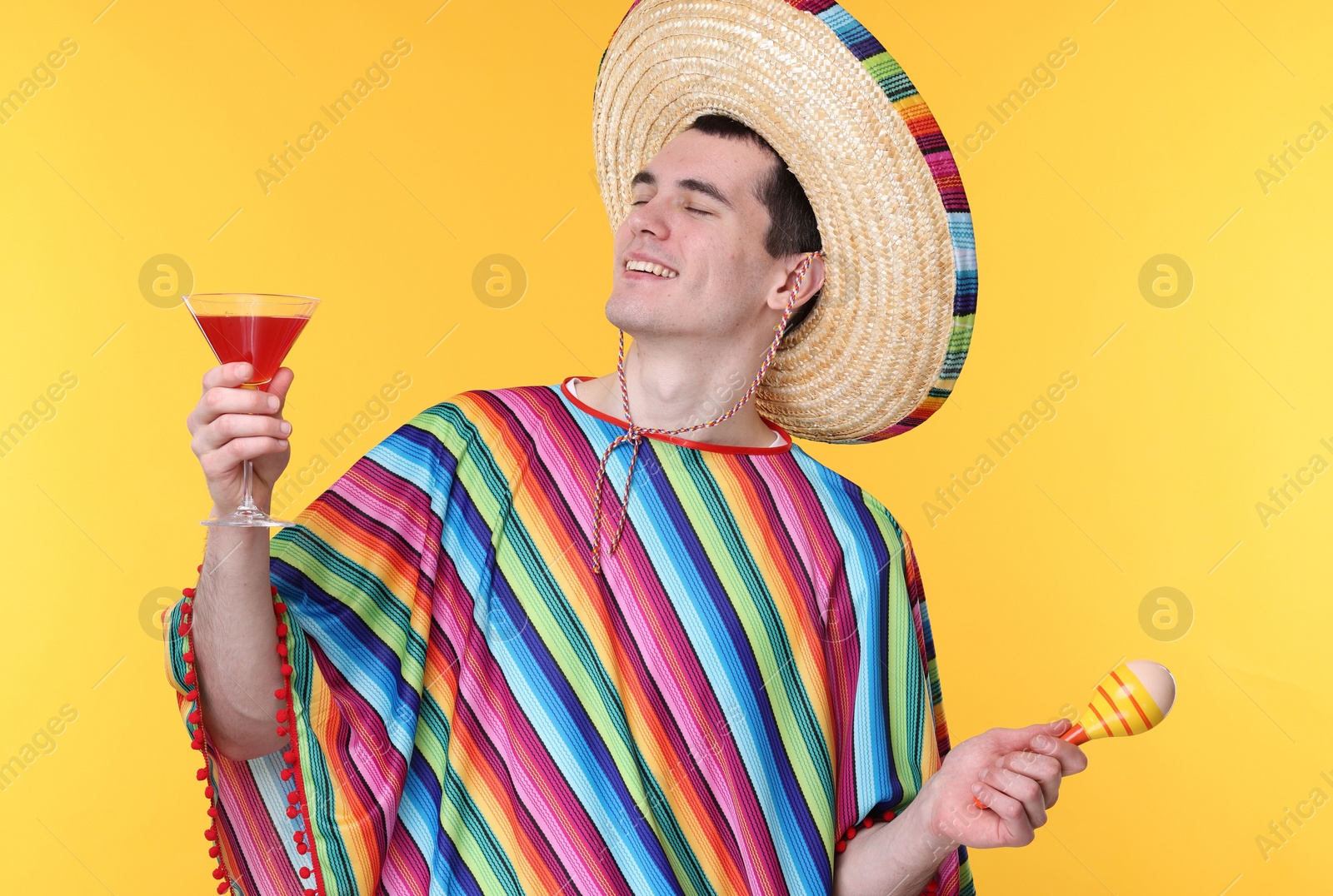 Photo of Young man in Mexican sombrero hat and poncho with cocktail and maracas on yellow background