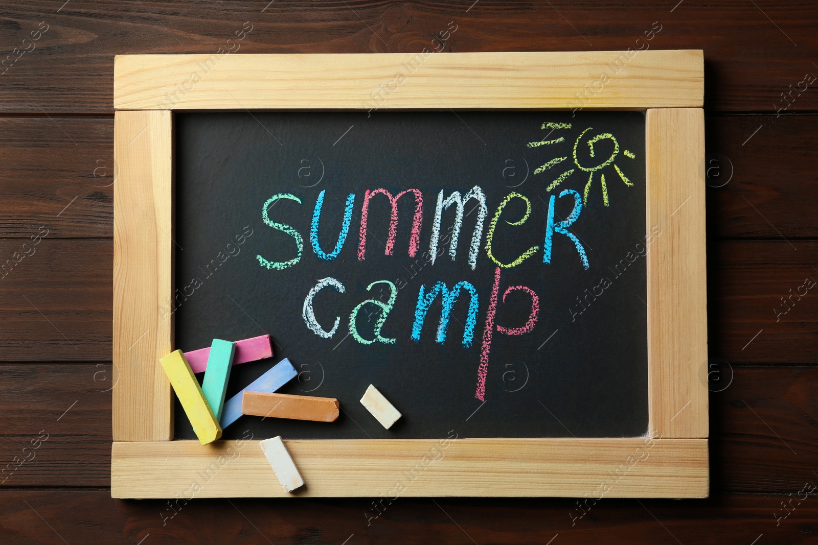 Photo of Small blackboard with text SUMMER CAMP, drawing and chalk sticks on wooden background, top view