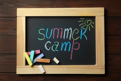 Photo of Small blackboard with text SUMMER CAMP, drawing and chalk sticks on wooden background, top view