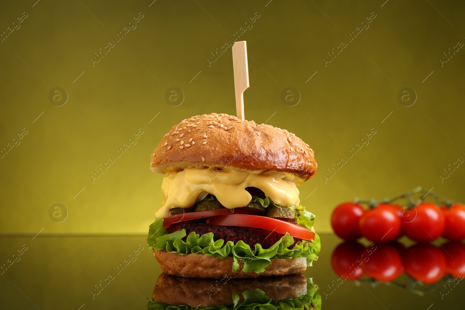 Photo of Delicious vegetarian burger and tomatoes on mirror surface against olive background