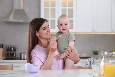 Happy young woman feeding her cute little baby at table in kitchen