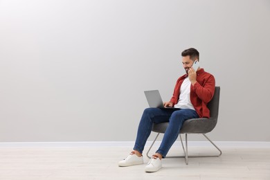 Photo of Handsome man with laptop talking on smartphone while sitting in armchair near light grey wall indoors, space for text