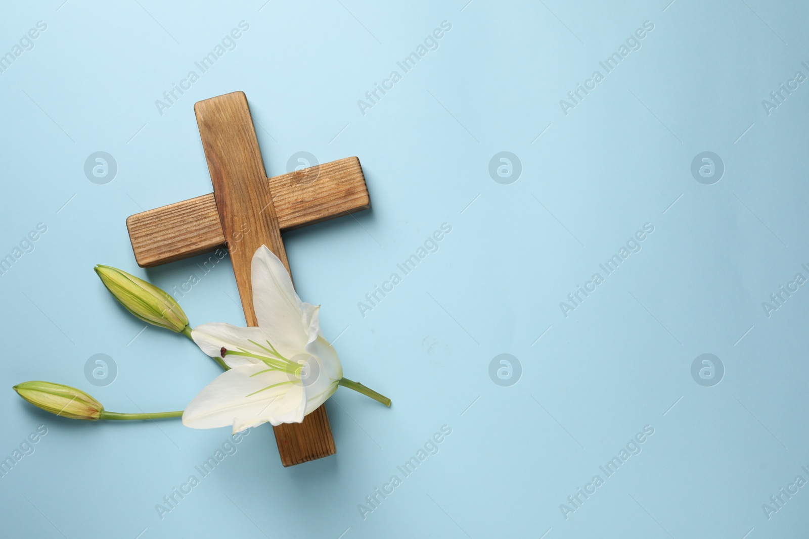 Photo of Wooden cross and lily flowers on light blue background, top view with space for text. Easter attributes