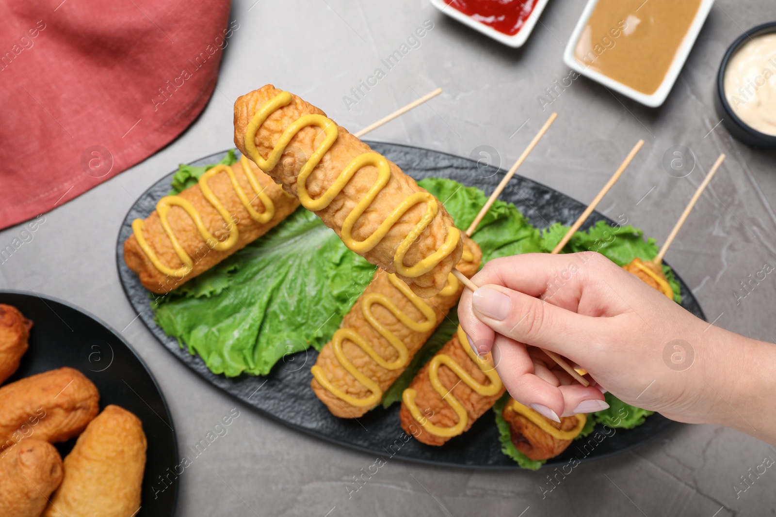 Photo of Woman holding delicious corn dog with mustard at grey table, top view
