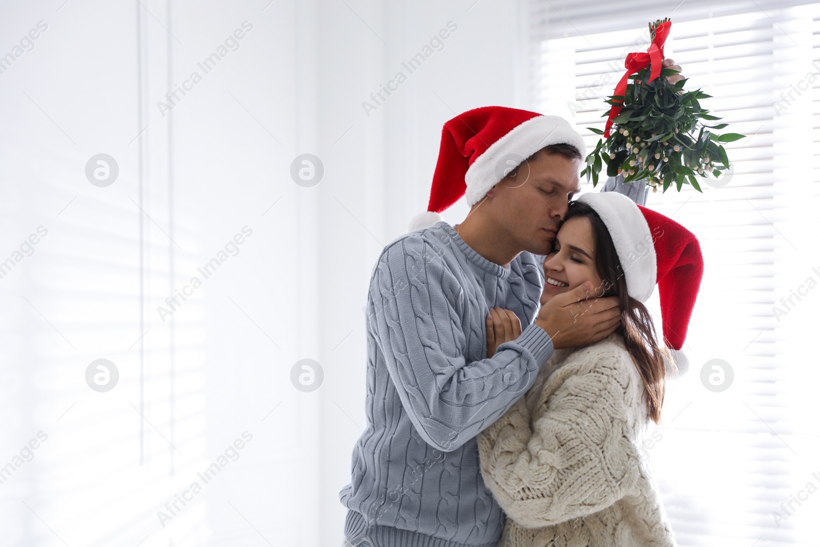 Photo of Happy man kissing his girlfriend under mistletoe bunch at home, space for text