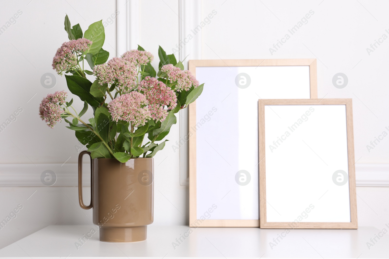 Photo of Stylish ceramic vase with beautiful flowers and blank frames on table near white wall