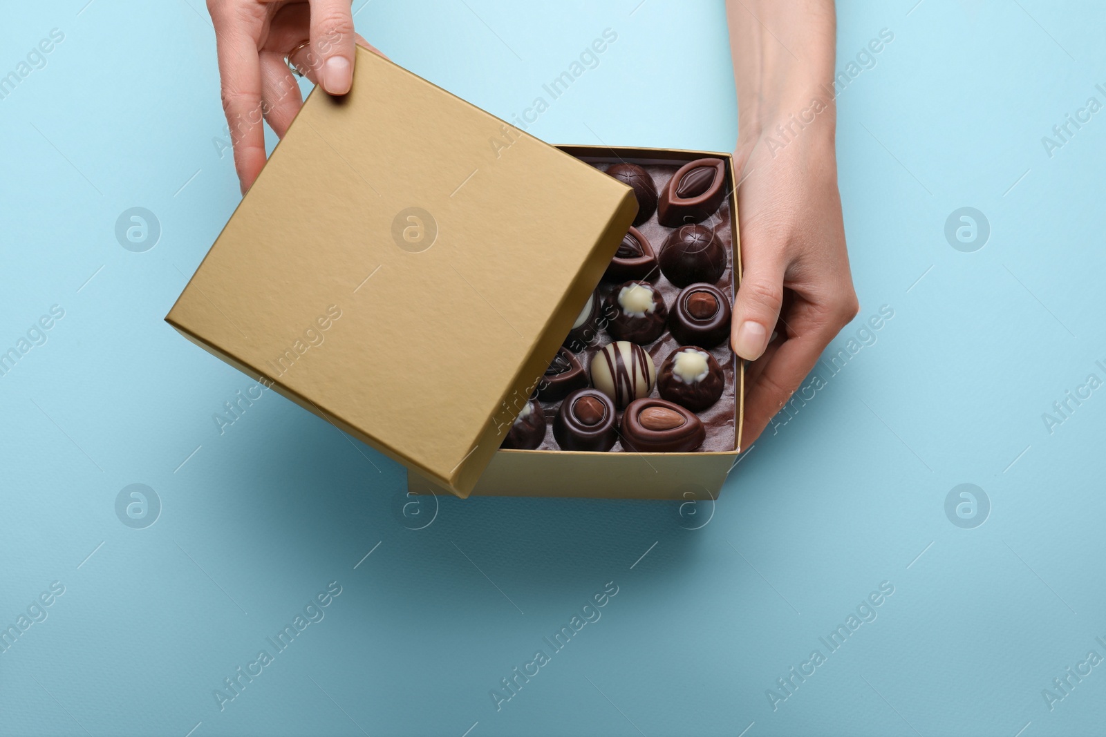 Photo of Woman with open box of delicious chocolate candies on light blue background, top view