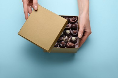 Photo of Woman with open box of delicious chocolate candies on light blue background, top view