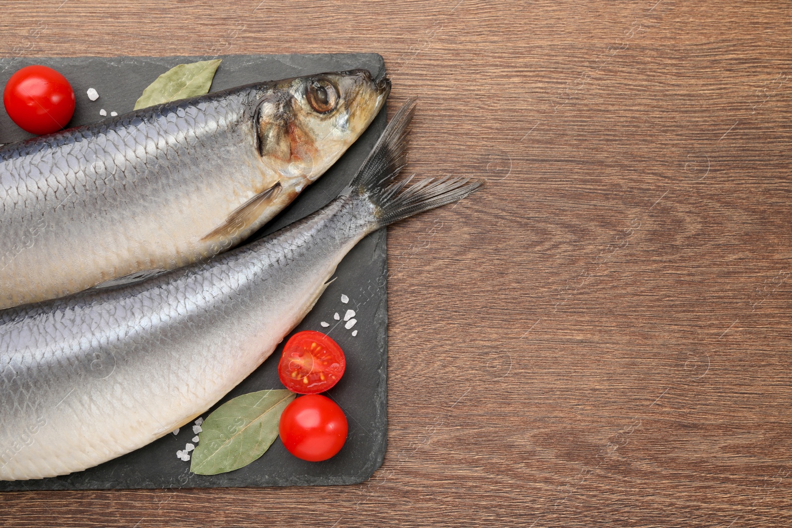 Photo of Slate plate with salted herrings, bay leaves and cherry tomatoes on wooden table, top view. Space for text