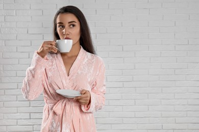 Photo of Young woman in bathrobe with cup of coffee near white brick wall. Space for text