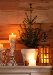Small potted fir, candles and decor elements on white mantel near wooden wall