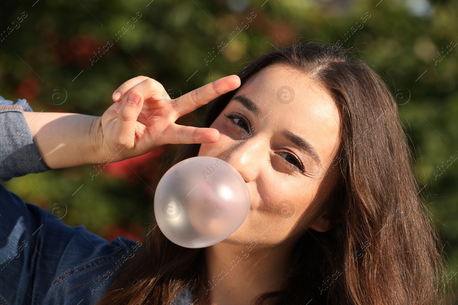 Photo of Beautiful young woman blowing bubble gum and showing V sign outdoors