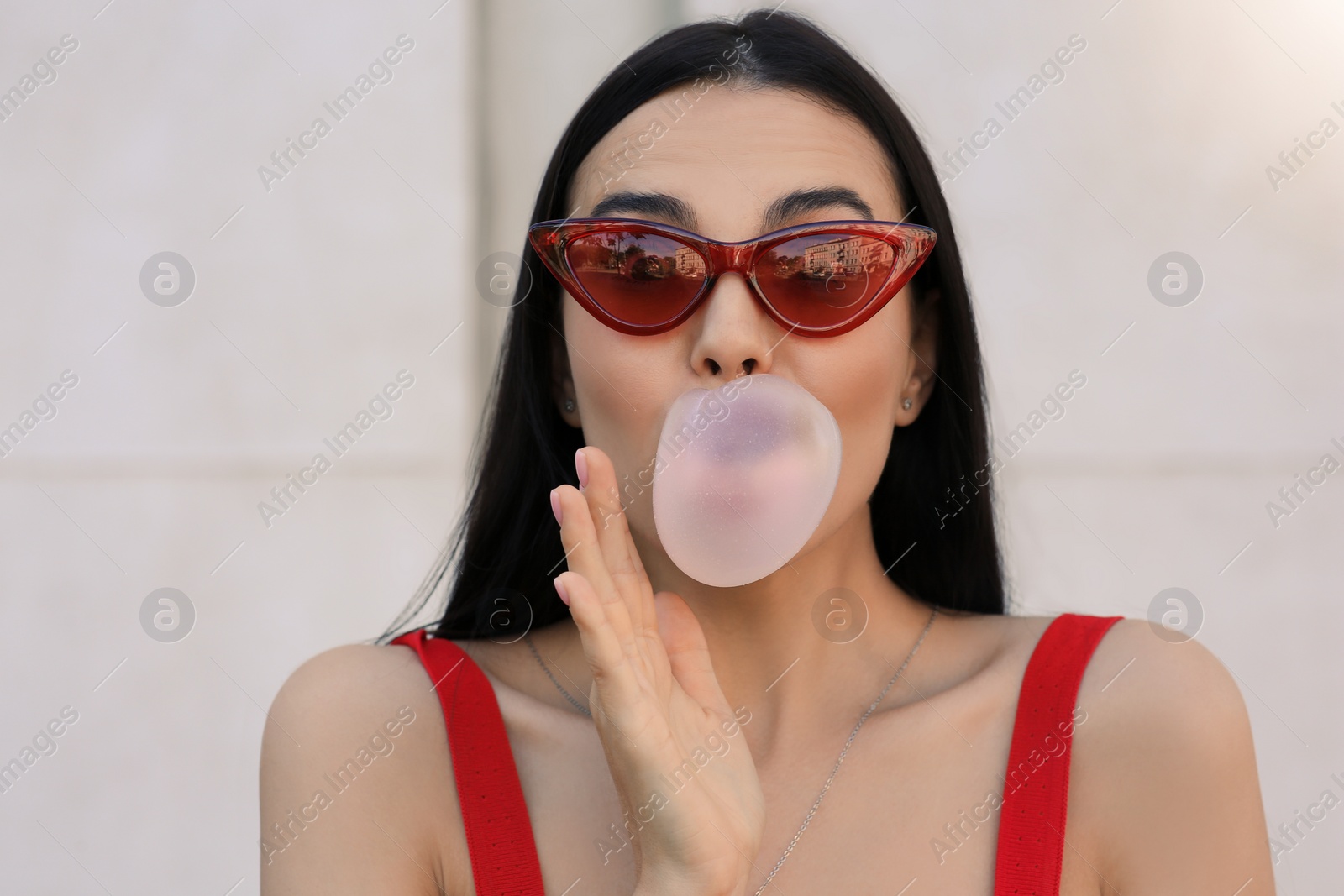 Photo of Beautiful woman in stylish sunglasses blowing gum near wall outdoors