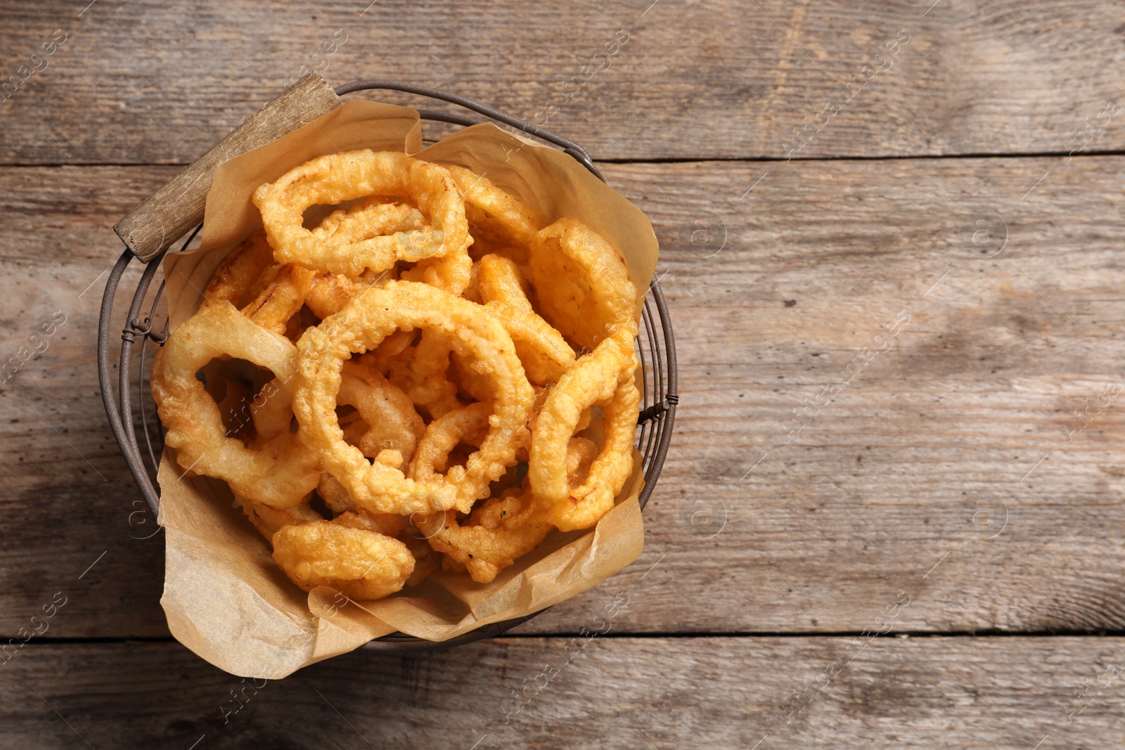 Photo of Homemade crunchy fried onion rings in wire basket on wooden background, top view. Space for text