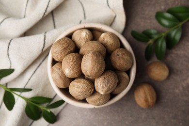 Photo of Whole nutmegs in bowl and green branches on brown table, flat lay