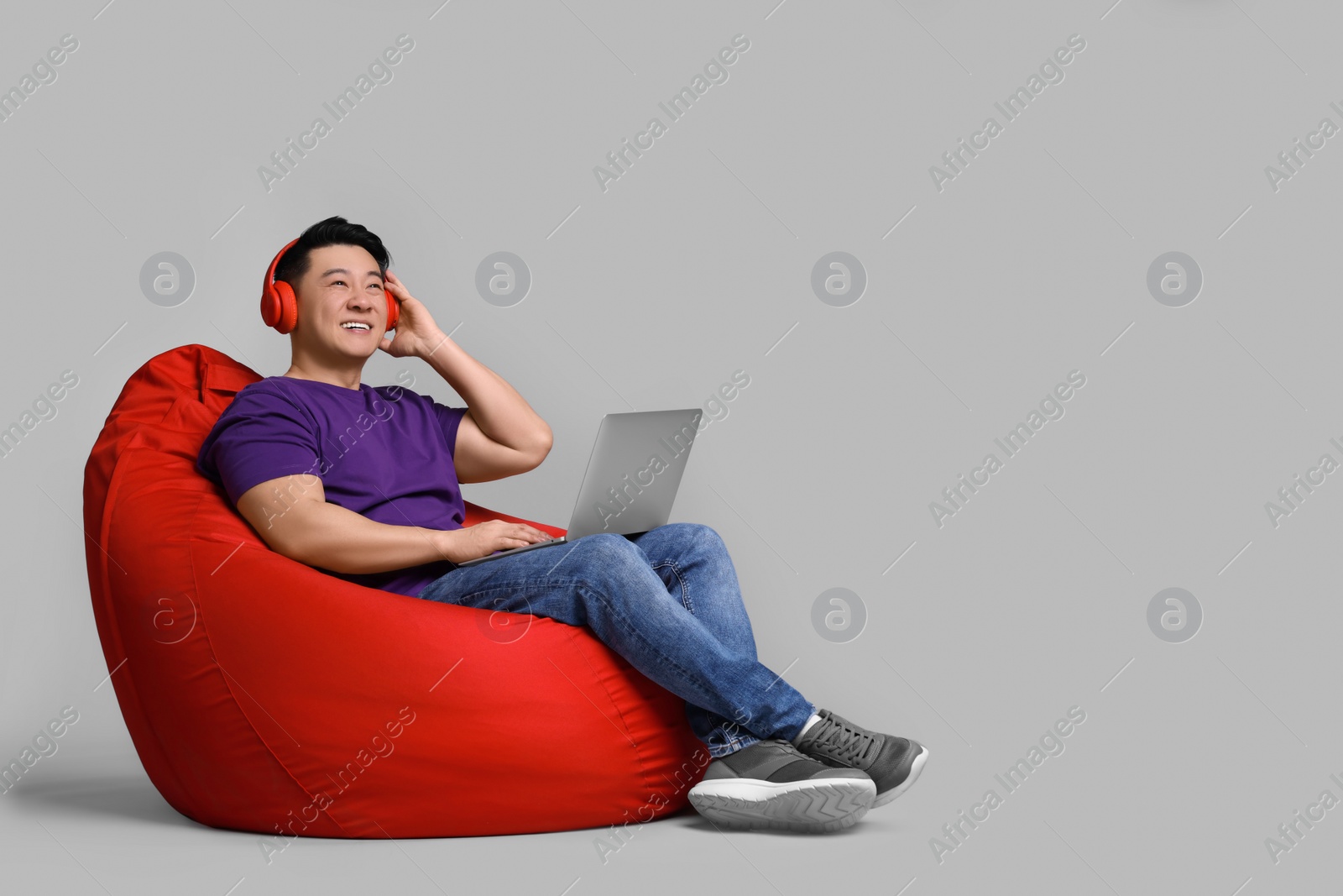 Photo of Smiling man with laptop and headphones sitting in beanbag chair against grey background