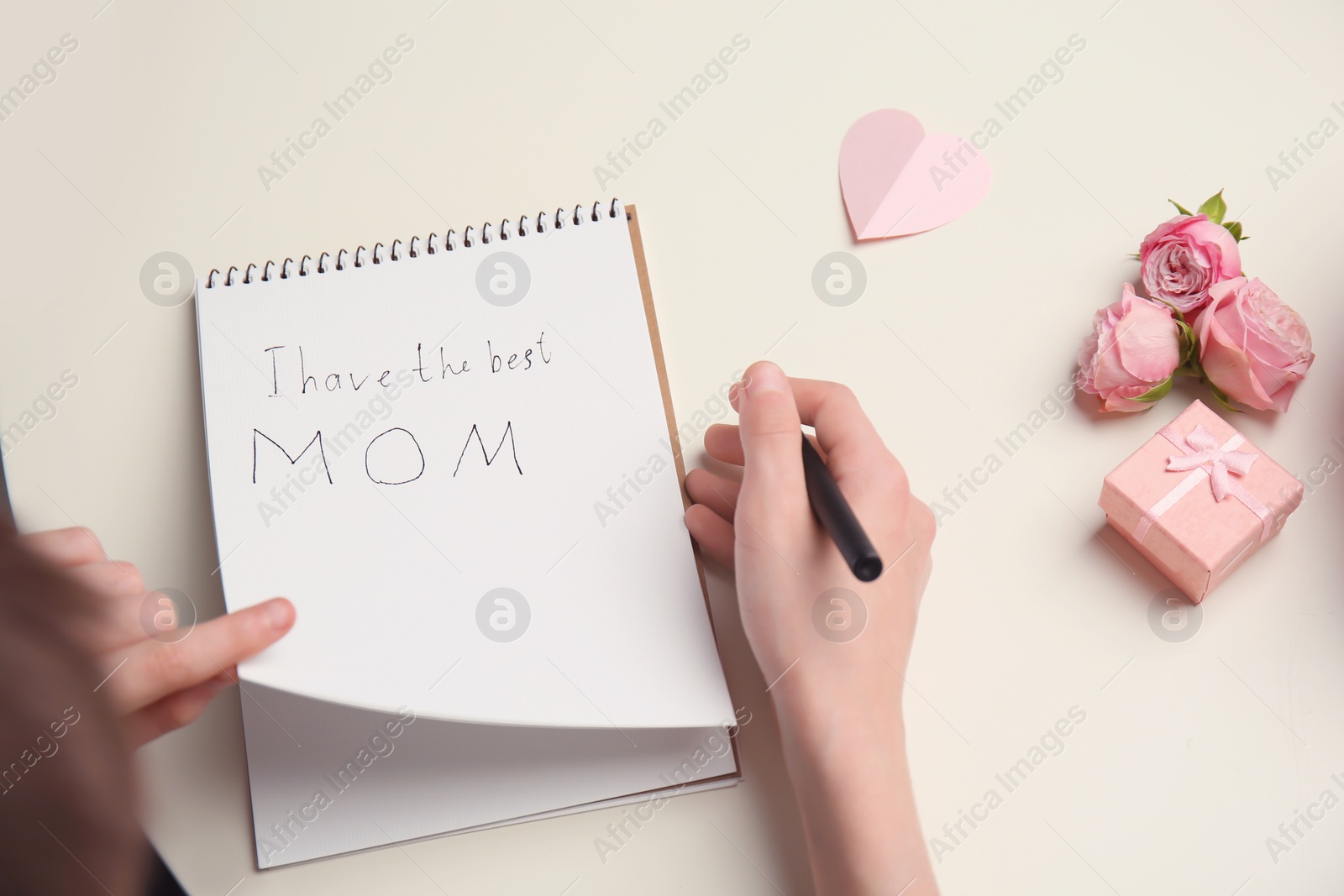 Photo of Little child writing congratulation for Mother's Day in notebook on light background, top view