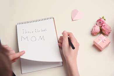 Photo of Little child writing congratulation for Mother's Day in notebook on light background, top view