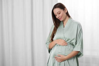 Photo of Beautiful pregnant woman in green dressing gown near window indoors, space for text