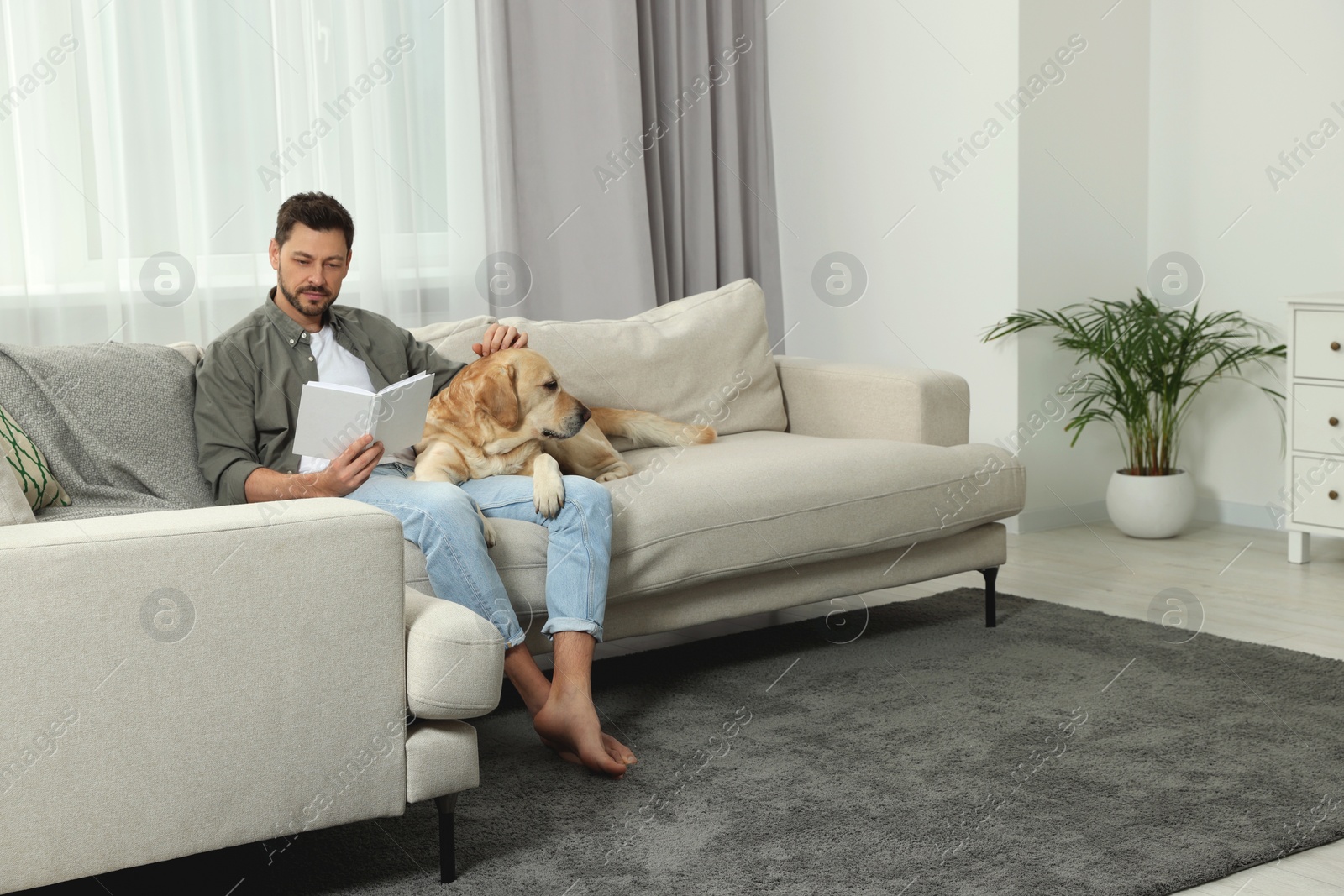 Photo of Man reading book on sofa near his cute Labrador Retriever at home. Space for text