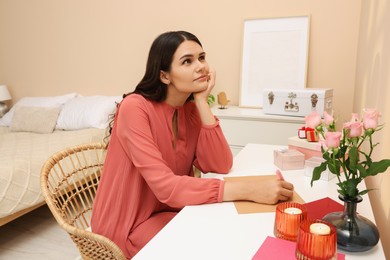 Photo of Young woman writing message in greeting card at table indoors