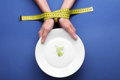 Woman holding plate with lettuce leaf in hands tied with measuring tape on blue background, top view. Diet concept