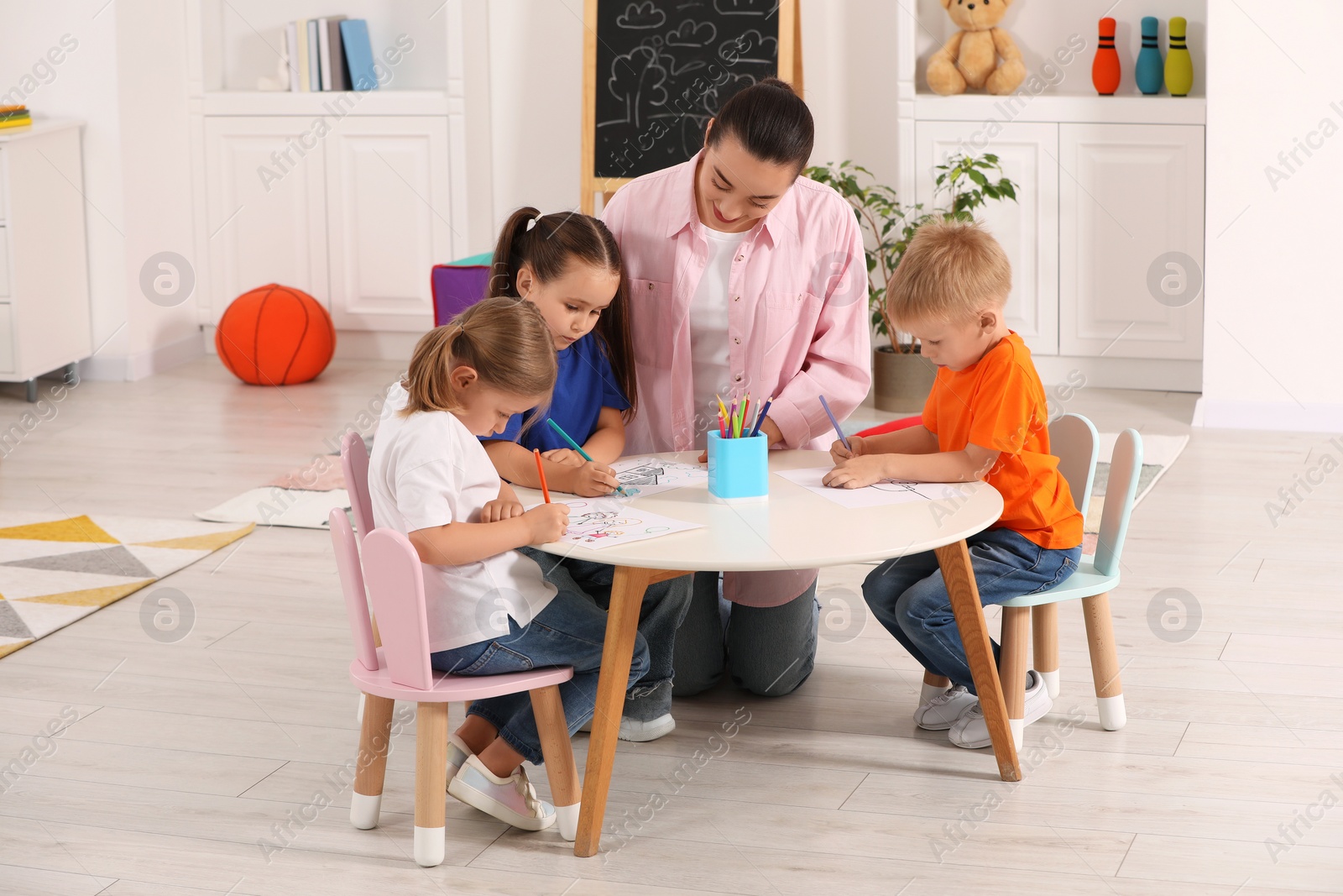 Photo of Nursery teacher and group of cute little children drawing at desk in kindergarten. Playtime activities