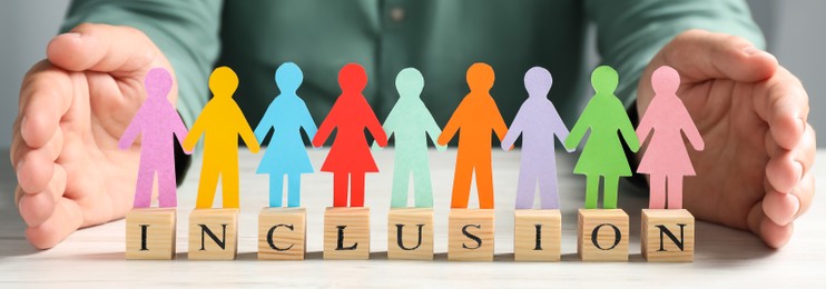 Image of Woman protecting colorful pawns and wooden cubes with word Inclusion at white table, closeup. Banner design