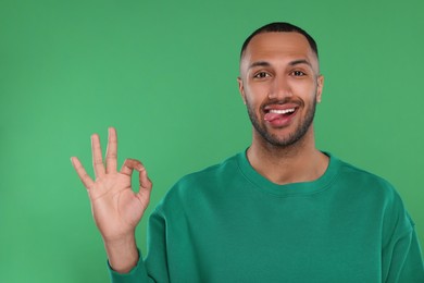Photo of Happy young man showing his tongue and OK gesture on green background