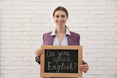 Teacher holding small chalkboard with inscription Do You Speak English? near white brick wall