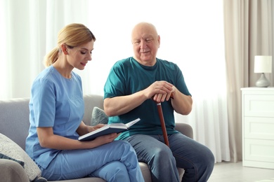 Nurse reading book to elderly man indoors. Assisting senior people