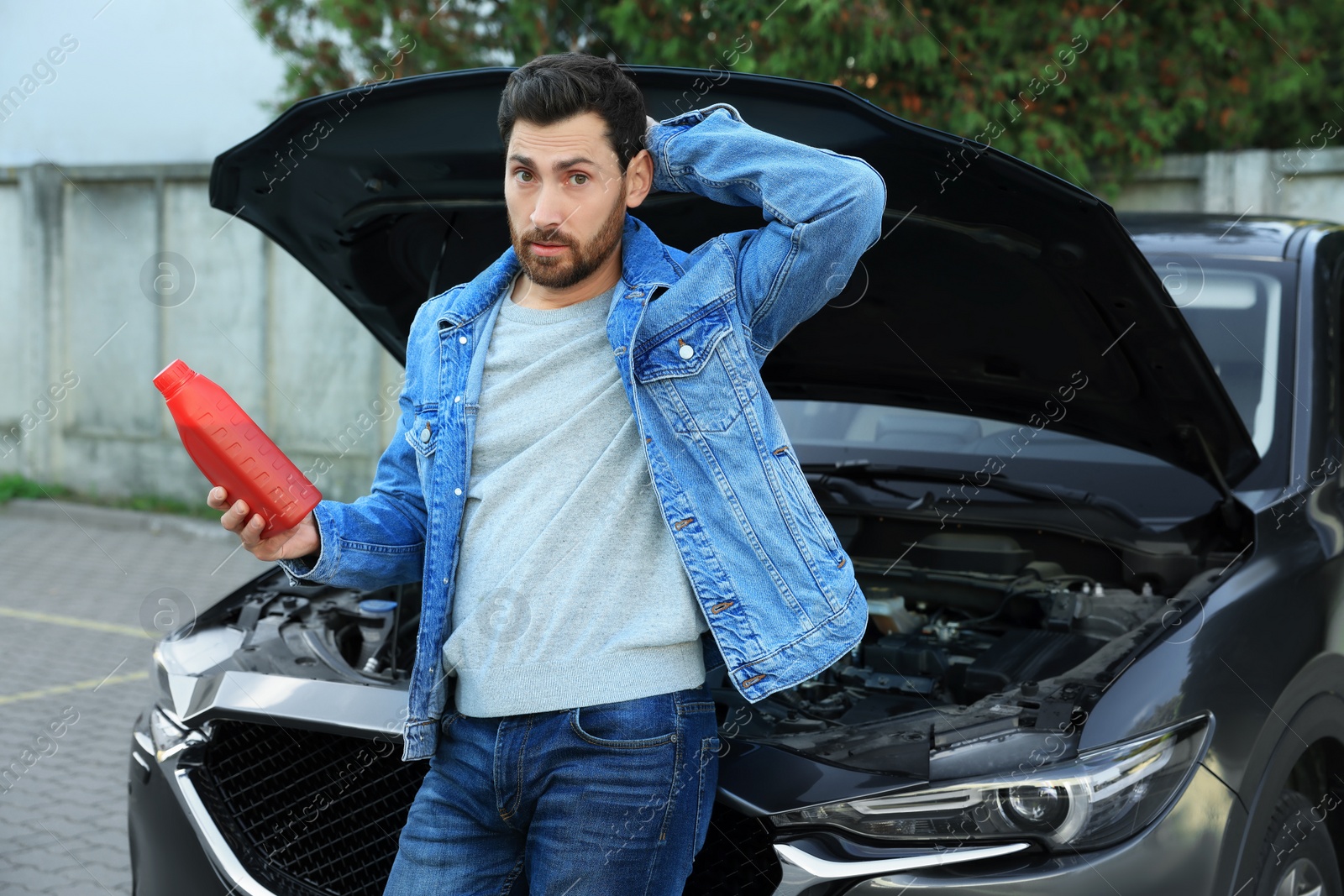 Photo of Puzzled man holding red container of motor oil near car outdoors