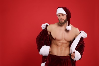 Muscular young man in Santa costume on red background, space for text