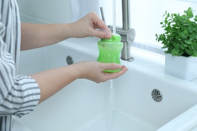 Photo of Woman washing baby bottle under stream of water in kitchen, closeup