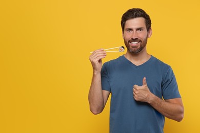 Photo of Happy man holding sushi roll with chopsticks and showing thumbs up on orange background. Space for text