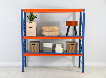 Photo of Metal shelving unit with different household stuff near light wall indoors