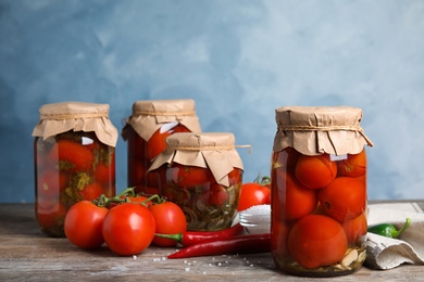 Pickled tomatoes in glass jars and products on wooden table against blue background