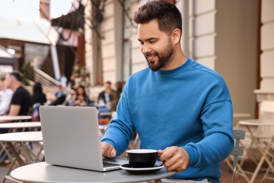 Handsome young man with cup of drink working on laptop at table in outdoor cafe