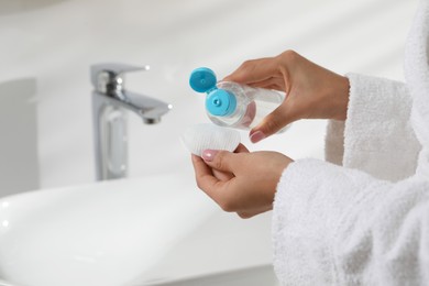 Photo of Woman pouring makeup remover from bottle onto cotton pad near sink indoors, closeup