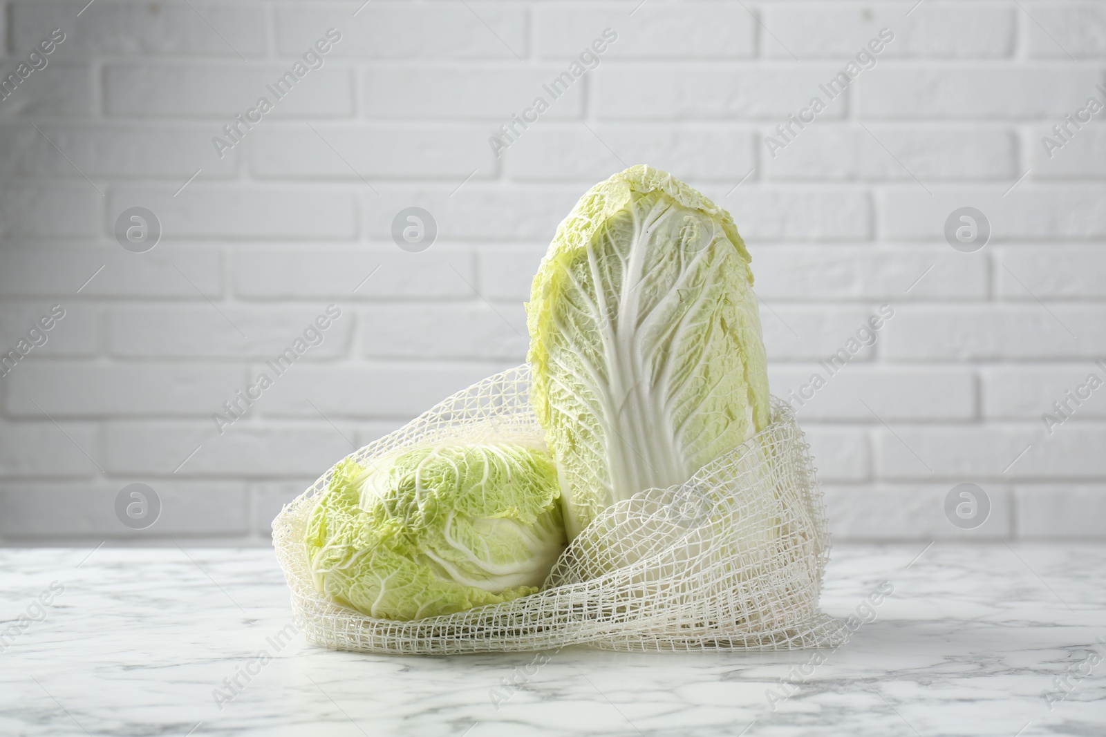 Photo of Fresh Chinese cabbages in string bag on white marble table near brick wall