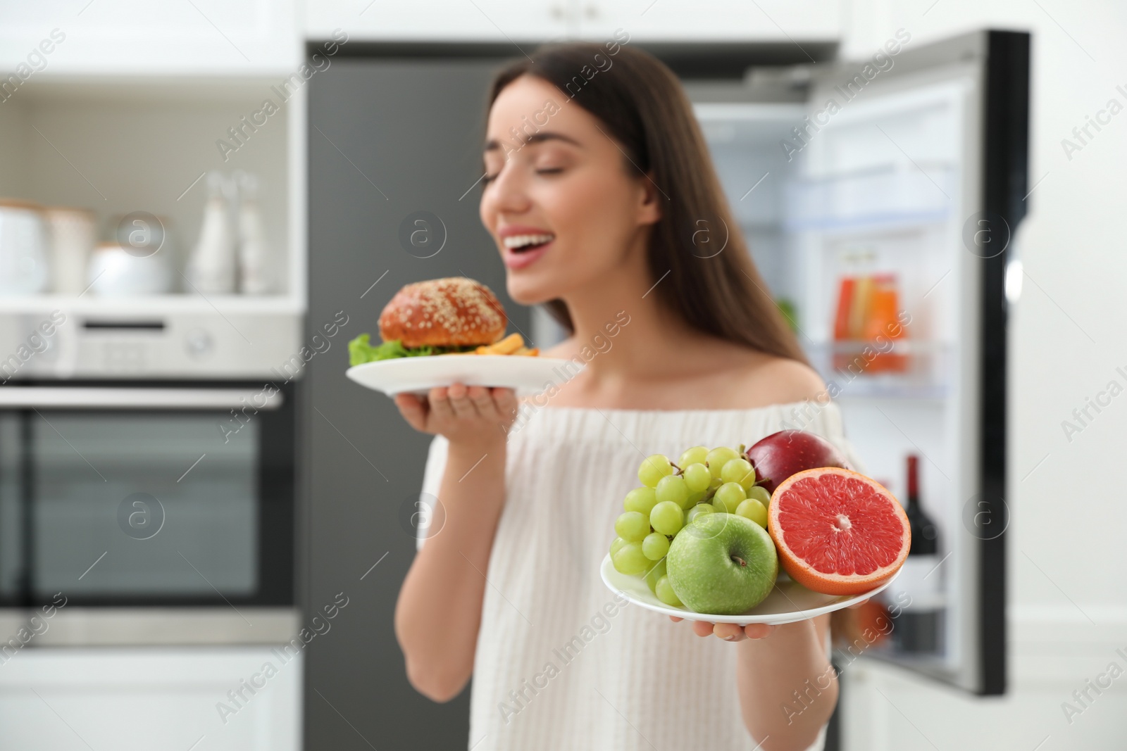 Photo of Woman choosing between fruits and burger with French fries near refrigerator in kitchen, focus on food