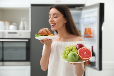 Photo of Woman choosing between fruits and burger with French fries near refrigerator in kitchen, focus on food